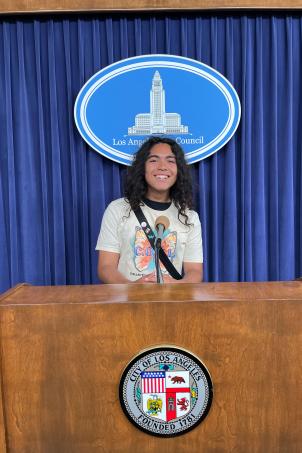 Medium shot of a high-school age kid with a medium skin tone and long hair standing indoors behind a wooden lectern with the City of Los Angeles seal on it and a blue curtain with the Los Angeles City Council logo on it.