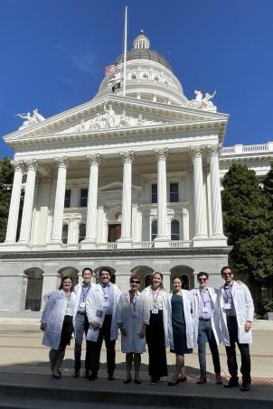 Eight medical residents wearing white lab coats smile as they stand in front of the California State Capitol in Sacramento