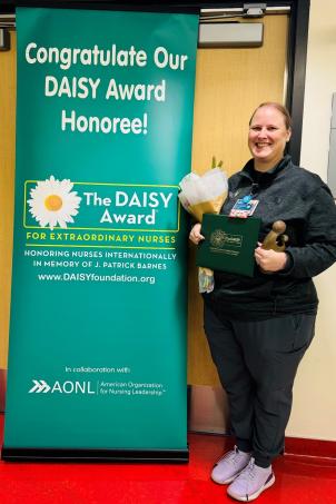 Female nurse with light skin tone and blonde hair smiles as she poses with their DAISY Award next to a DAISY Award poster