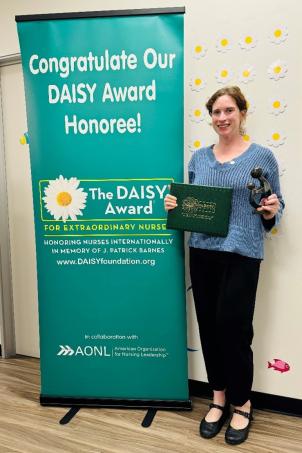 Female nurse with light skin tone and blonde hair smiles as she poses with their DAISY Award next to a DAISY Award poster