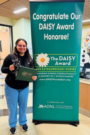 Female nurse with medium skin tone, brown hair and glasses smiles as they pose with their DAISY Award next to a DAISY Award poster