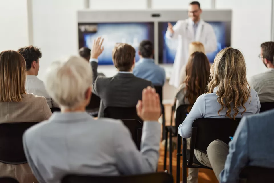 Man with light skin tone wearing lab coat and glasses instructs group of professionally dressed adult students in classroom setting