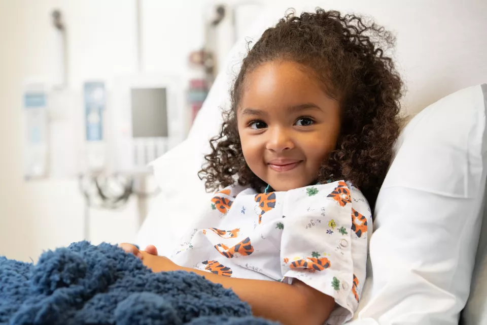 Little girl with medium skin tone and wearing tiger printed pajamas smiles as she lies in a hospital bed