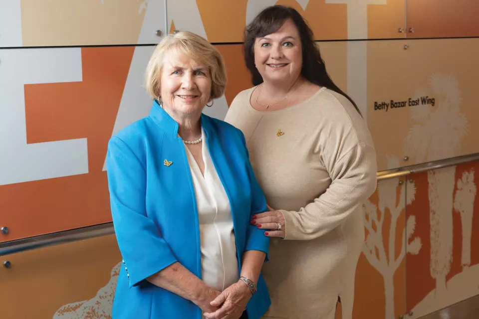 A woman with light skin tone, short blonde hair wearing a light blue jacket standing next to a woman with long dark hair wearing a beige dress, in a hospital corridor.