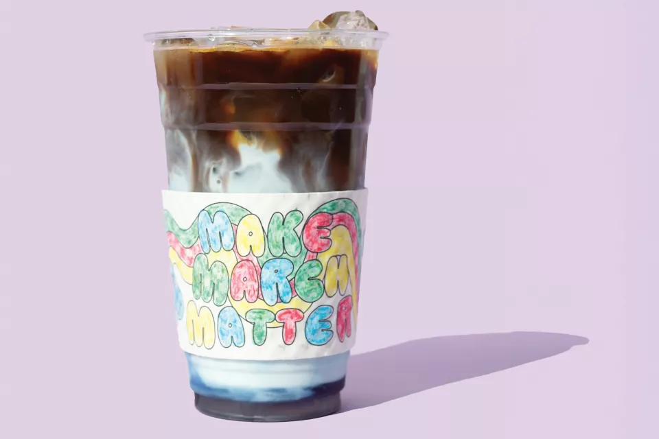 A cup of iced coffee with milk in a cup sleeve with the words Make March Matter in colorful lettering placed on a lavender background