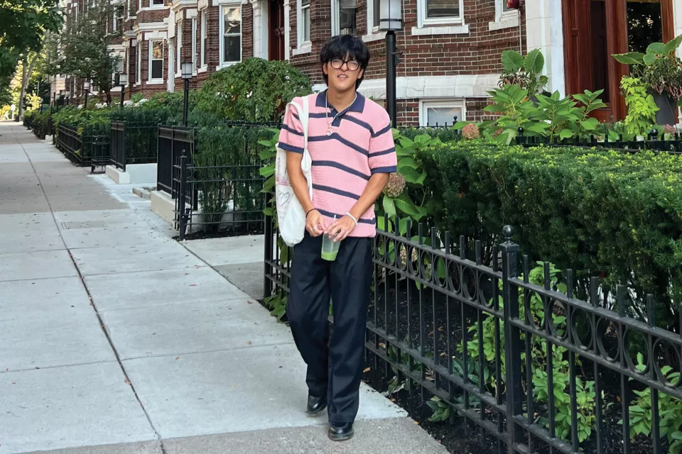 Teenage boy with medium skin tone and dark hair wearing a black and pink short sleeved collared shirt and long black pants, standing outside near a brick building.