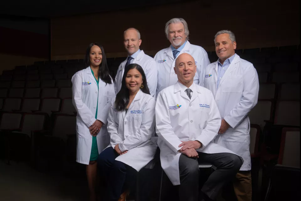 Seven physicians wearing white lab coats arranged in two row against a darkened background