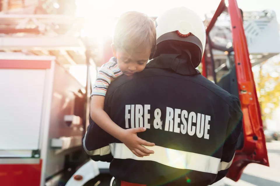 First responder carries a young boy with light skin tone and brown hair toward a fire truck