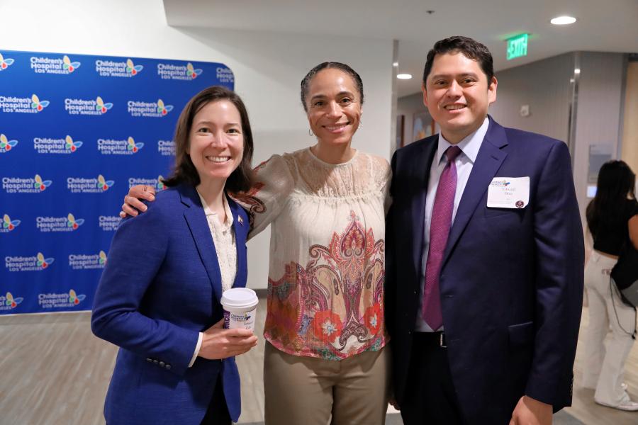 Close shot of two women with a medium-light skin tone standing with a man with a medium-light skin tone, all smiling and wearing formal attire in a room with the Children’s Hospital Los Angeles logo on a blue background behind them.