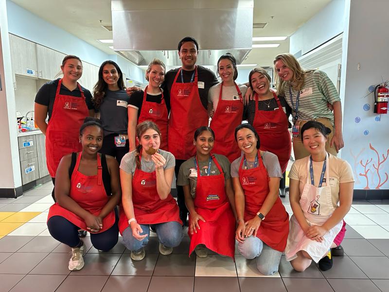 Twelve medical residents wearing bright aprons smile as they pose for a team photo in a kitchen setting