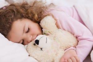 Little girl with light skin tone and curly brown hair hugs teddy bear as she sleeps