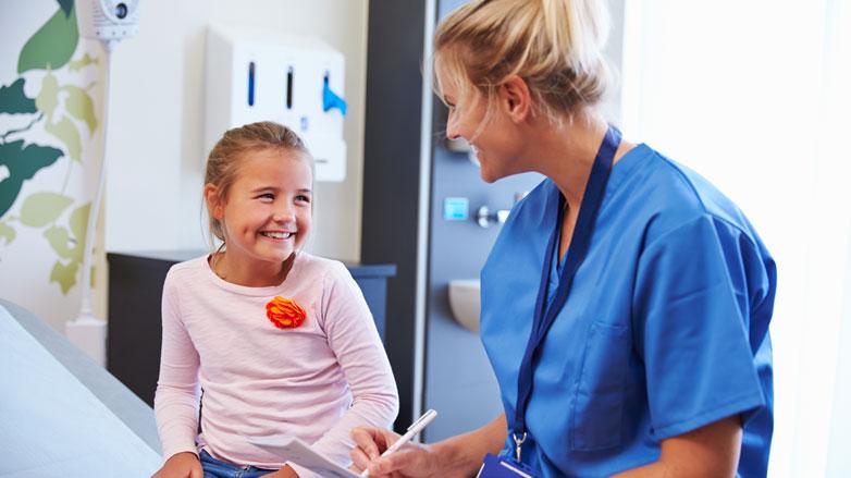 A young light skin-toned girl and a light skin-toned nurse in blue scrubs smile at each other